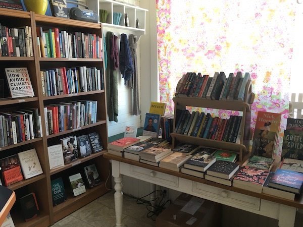 A photo inside Happenstance Books & Yarns taken by the front window with a pink, green, and yellow floral curtain covering it. There is a white wooden table by the window with books displayed on top and wooden bookshelves lining one side of the store with books on them, spines facing out.