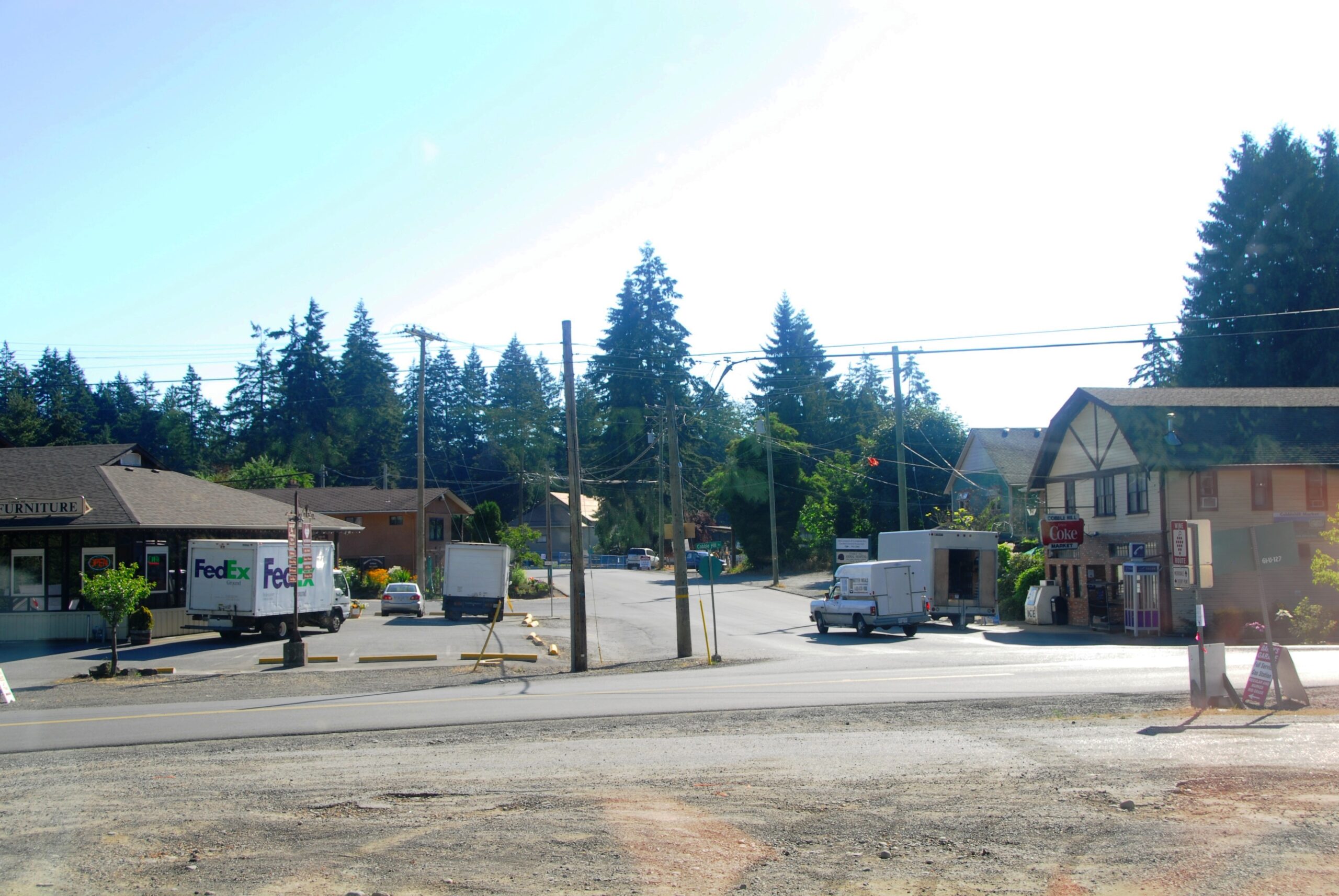A photo of Cobble Hill, BC. A T-intersection shows an old-fashioned corner store, a few cars and trucks driving about or parked, and coniferous trees fringing the down. The day is bright.