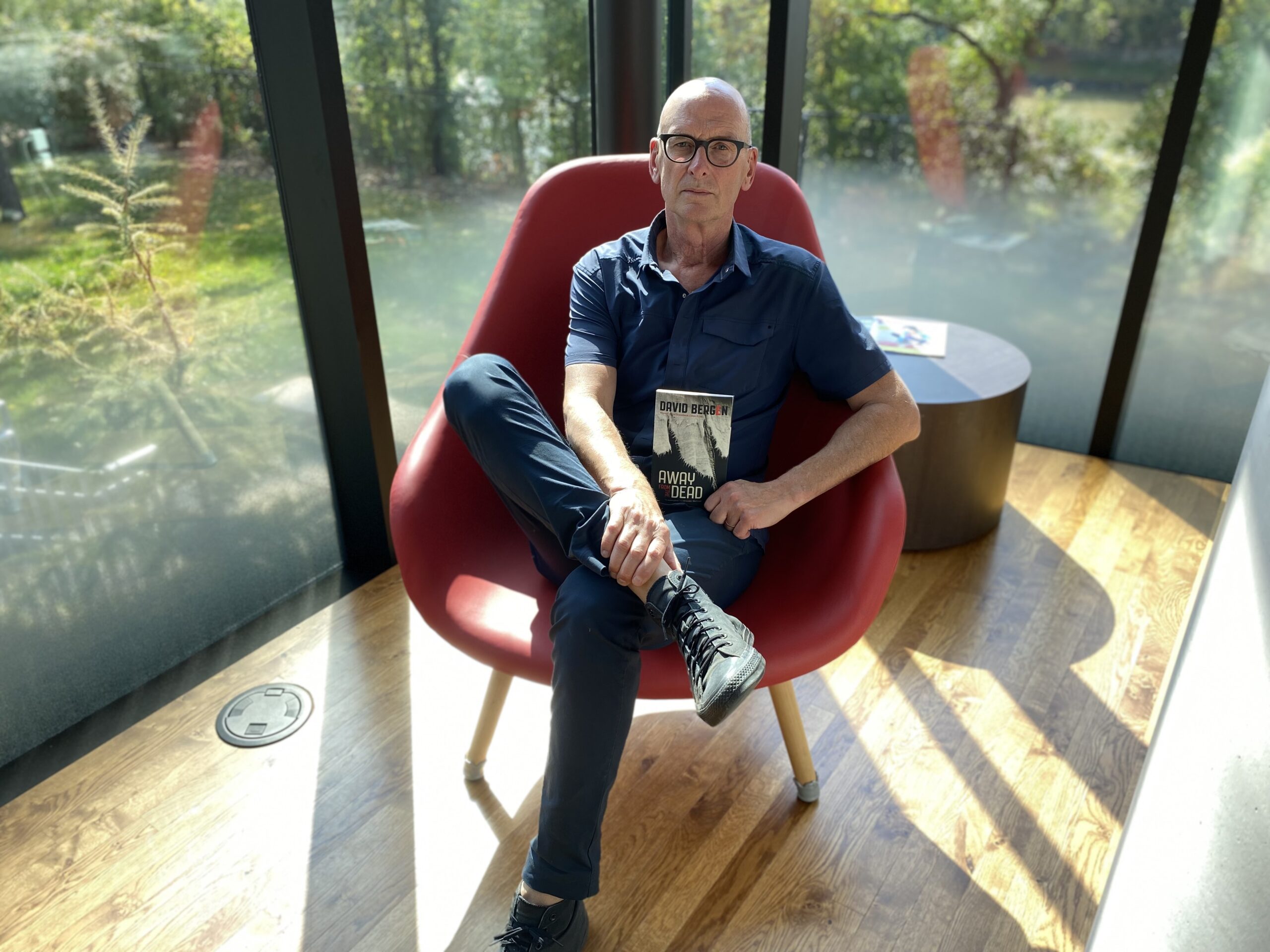A photo of author David Bergen holding a copy of his book Away from the Dead. He sits in a wide, comfortable red lounging chair in the corner of a glass-walled room, looking on to a bright, sunny green yard.