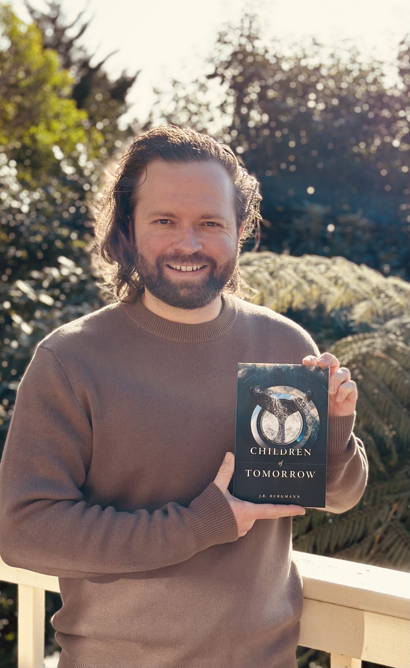 A photo of author J.R. Burgmann, a white man with a beard and shoulder-length hair, holding a copy of his book, Children of Tomorrow.