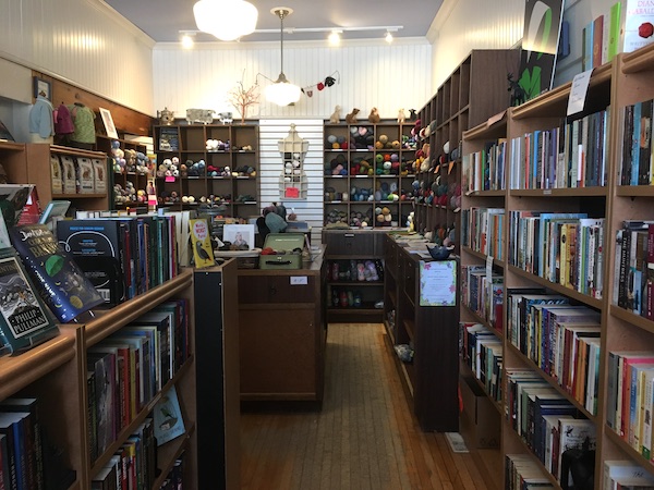 A photo inside Happenstance Books & Yarns that shows rows of wooden bookshelves with books on them, spines facing out. At the back of the shop are shelves with bundles of brightly-coloured yarn.