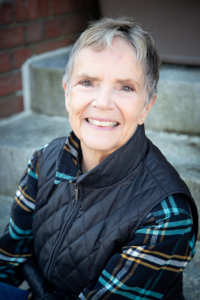 A photo of author Leslie Davidson. She is a white woman with short-cropped grey hair and a light puffer vest on over a plaid shirt. She smiles widely at the camera.