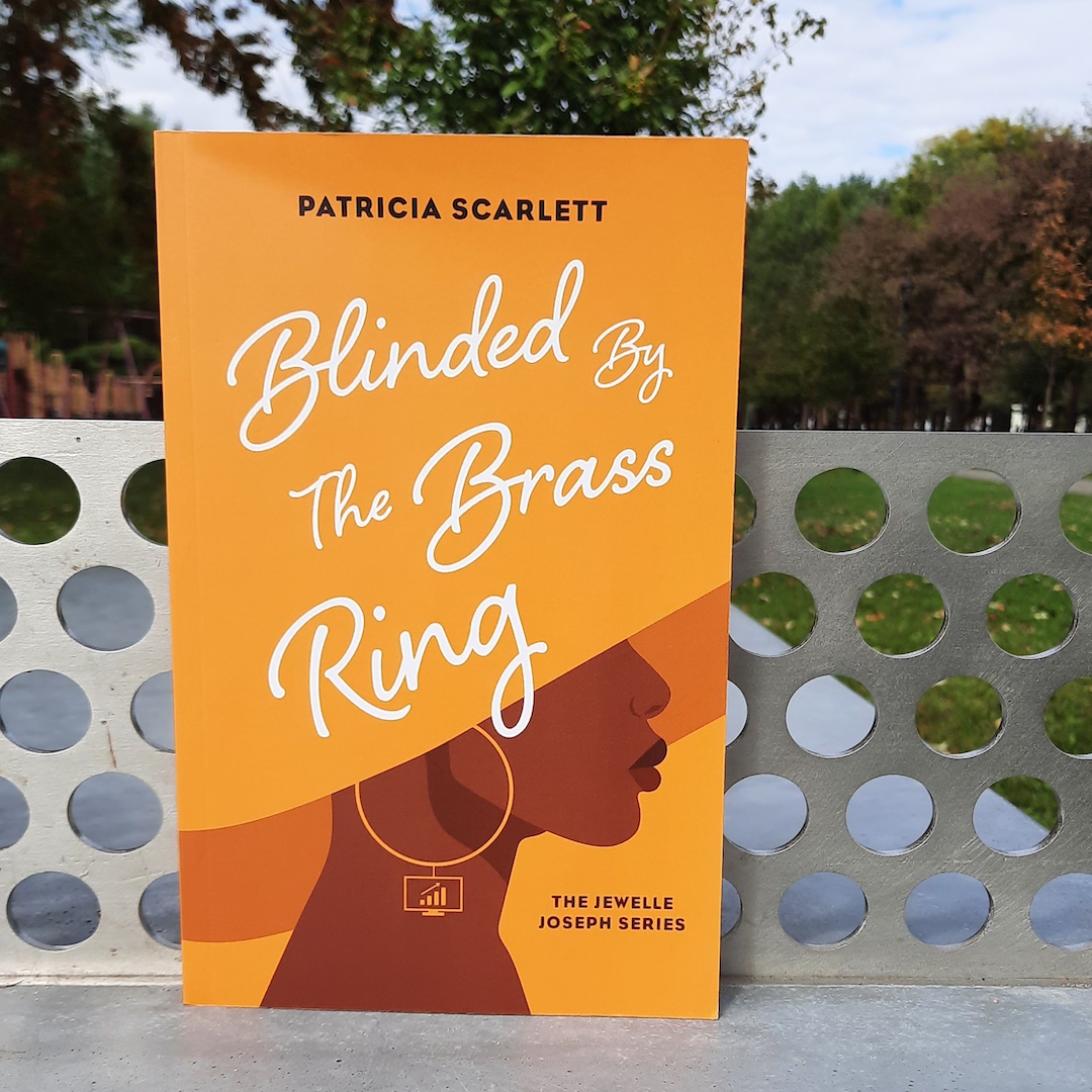 A copy of Blinded by the Brass Ring, standing in front of a perforated metal ledge, overlooking a park.