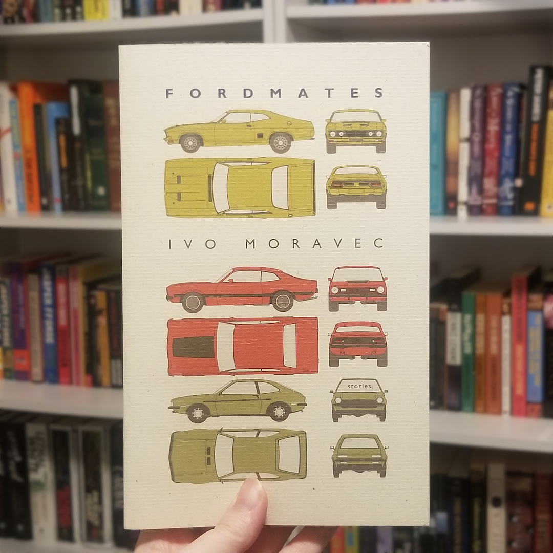 A light skin-toned hand holds up a copy of Fordmates by Ivo Moravec. The cover features illustrations of muscle cars at various angles. There is a bookshelf in the background.