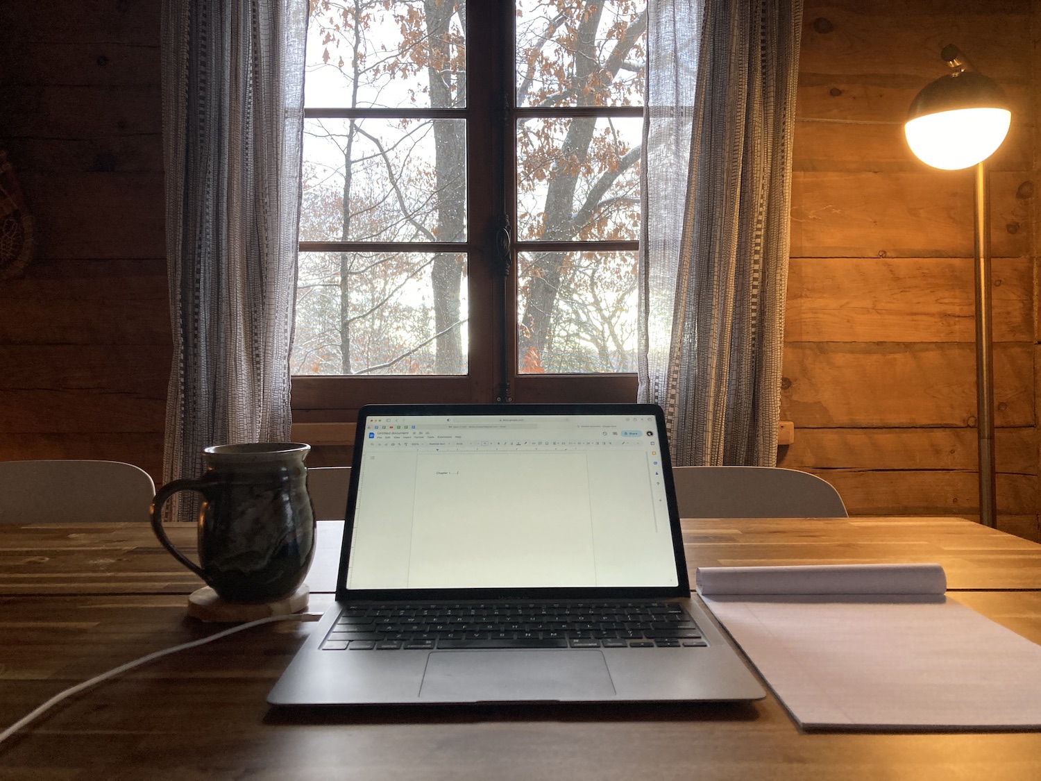 An open laptop sits next to a mug and notepad, in front of a window with a large tree outside. The walls in the room are wooden, like in a cabin.