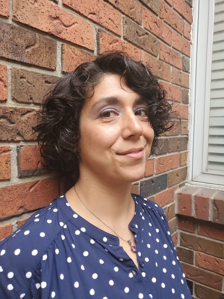 A photo of Kimia Eslah, an Iranian woman with chin-length curly hair and a polka dot top. She leans against a brick wall and smiles at the camera.