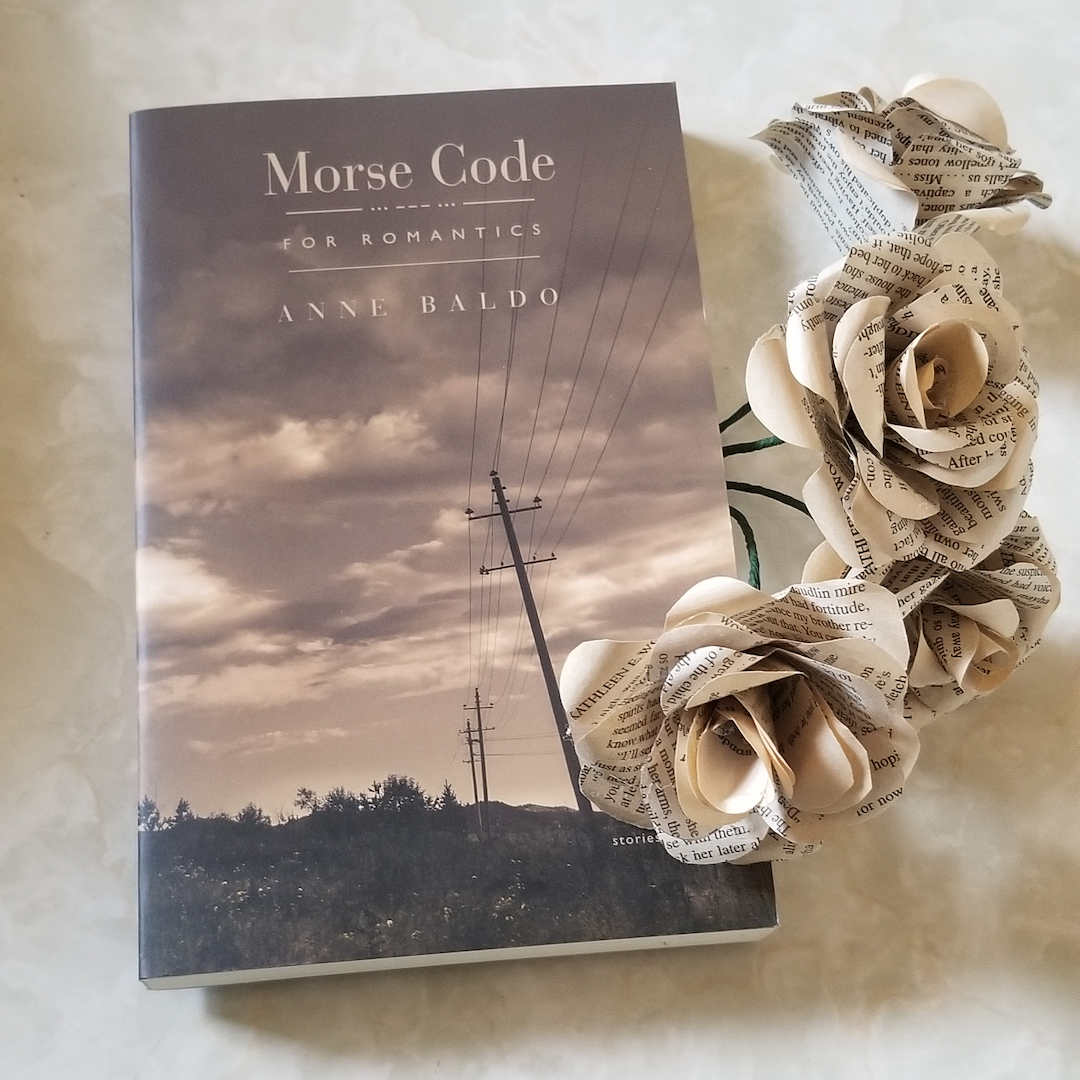 A photo of the book Morse Code for Romantics by Anne Baldo. The book lays next to roses made from the pages of books.
