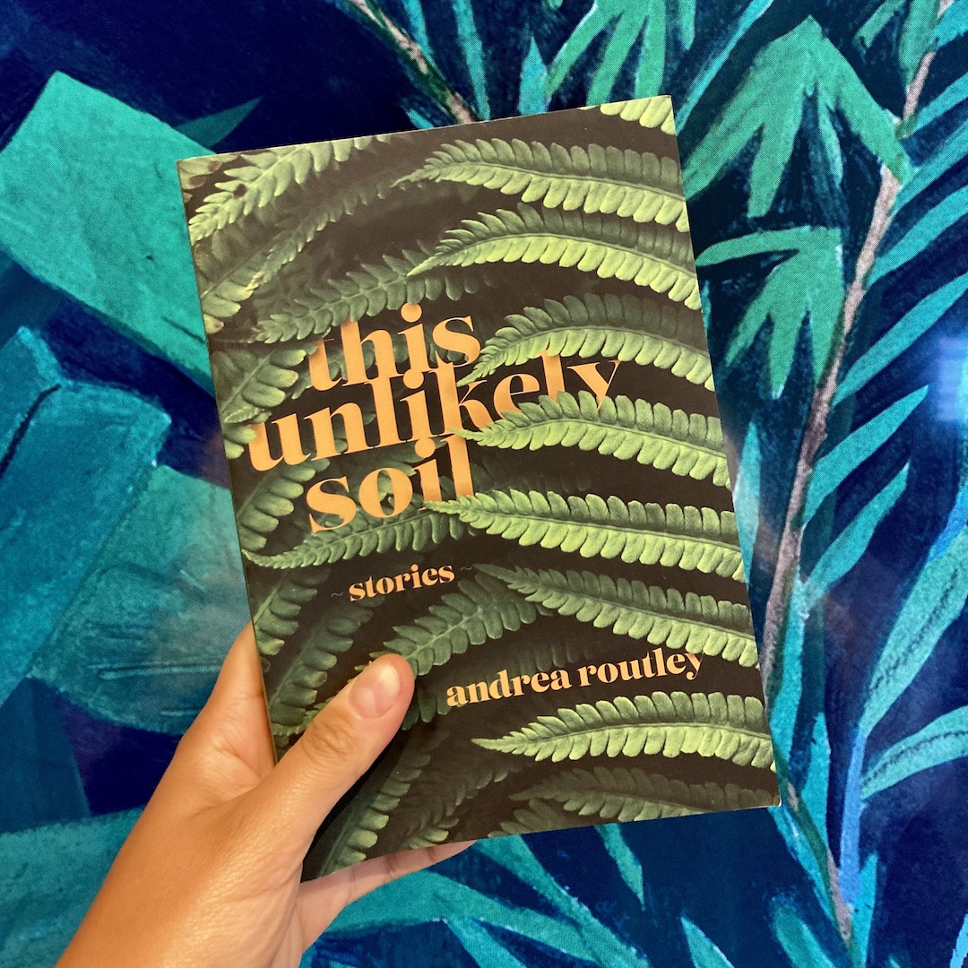A darker skin-toned hand holds up a copy of This Unlikely Soil by Andrea Routley, against a vibrant backdrop of tropical leaves.