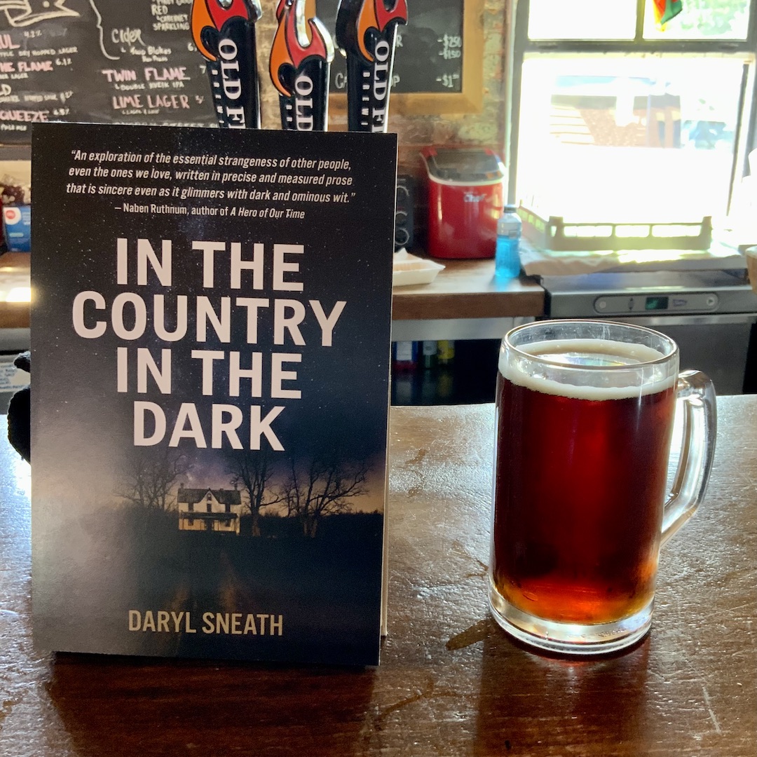 A copy of In the Country in the Dark sits next to a full pint of reddish beer, on a bartop.