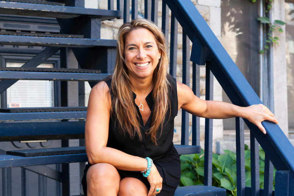 A photo of author Toula Drimonis. A white woman with long, dark blonde hair, she sits on an outdoor staircase and smiles at the camera.