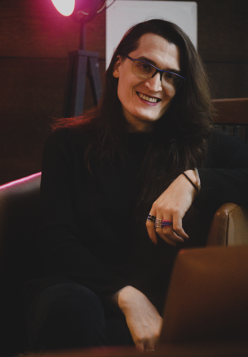 A photo of author AGA Wilmot, a nonbinary person with light skin tone and long, dark hair. They are sitting in a relaxed pose in a chair, and wear many rings on their fingers.