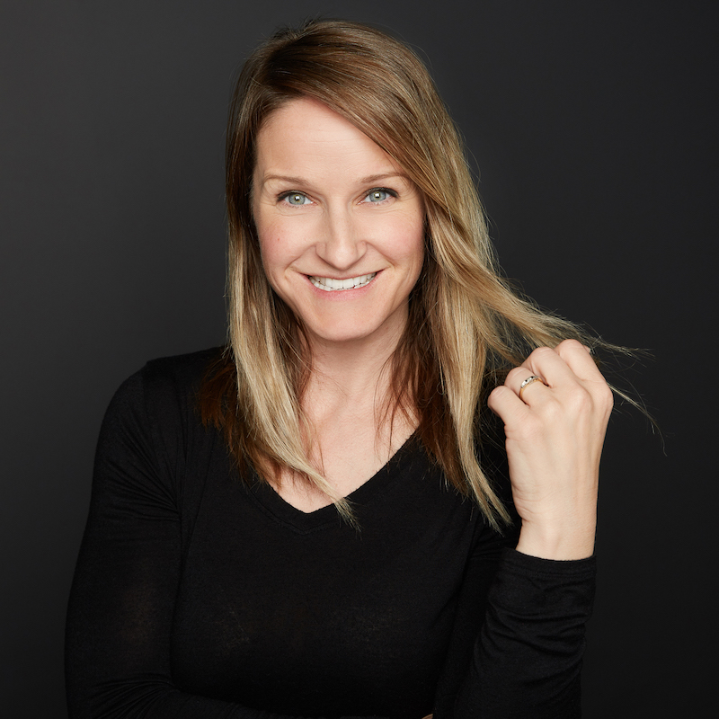 A photo of author Ali Bryan. A light-skinned woman with dark blonde hair, she beams at the camera and holds a strand of hair in her hand.
