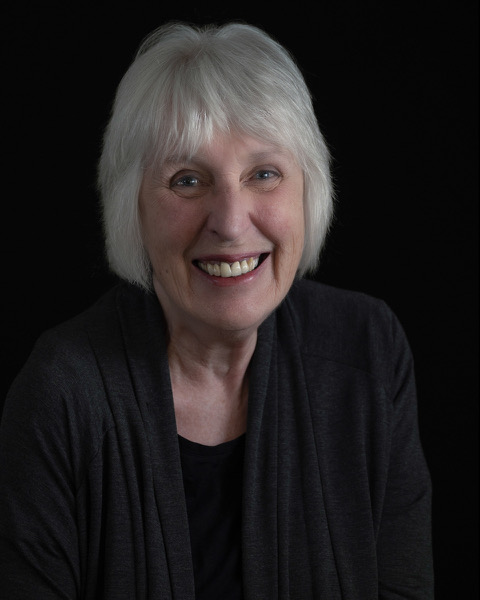A photo of writer Raye Anderson. She is a light-skinned woman with chin-length grey/white hair, and she wears a dark sweater.