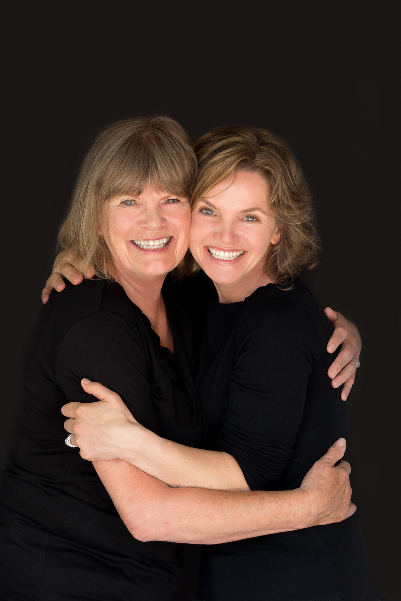 A photo of writers Alie and Hejsa Christensen. They are a mother and daughter, with similar dark blonde hair and wide smiles. They embrace each other and look toward the camera.