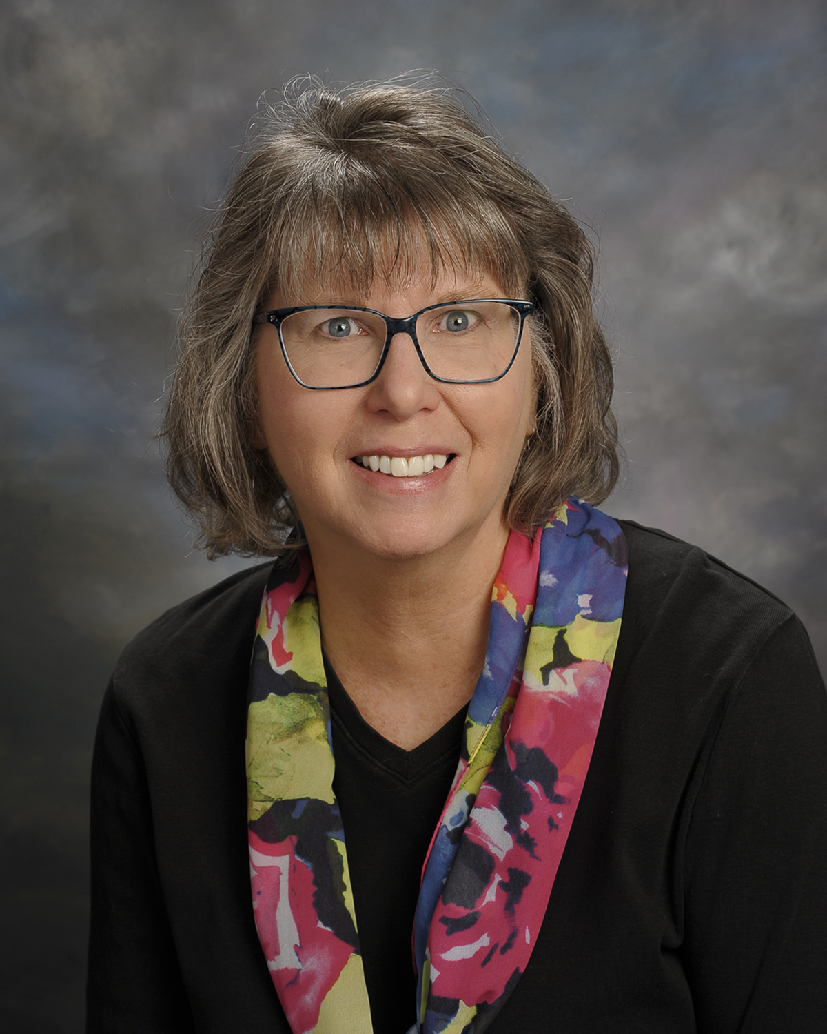 A photo of writer Marion Agnew, a light-skinned woman with short cut grey hair, wearing glasses and a bright patterned scarf.