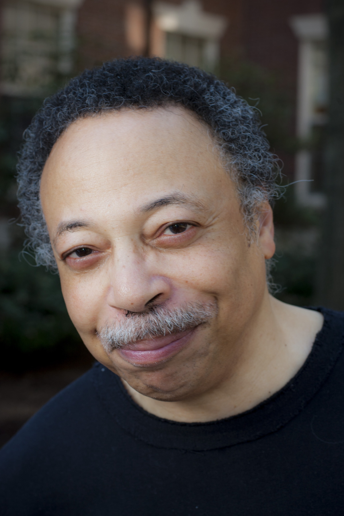 A photo of George Elliott Clarke. A Black man wearing a black shirt, he has a greying moustache and hair, and is smiling at the camera.