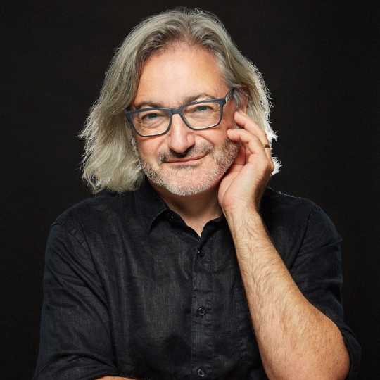 A photo of writer Gary Barwin. He is a light skin-toned man with chin-length grey hair and glasses, and he wears a black button down shirt against a black background.