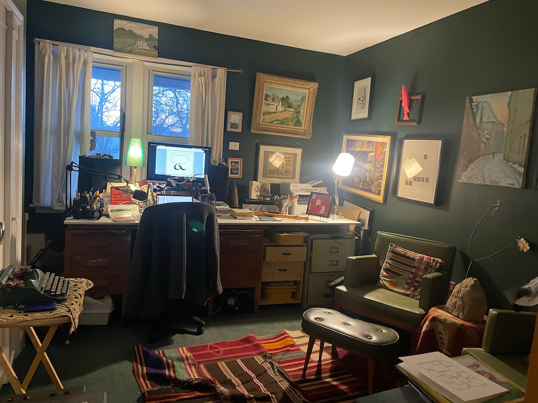 A photograph of Gary Barwin's writing space: a dark-painted, cozy office with a view of trees out the window. There is a long desk with a computer and various other tools and objects on it, an easy chair with a footrest, and a typewriter on a small table.