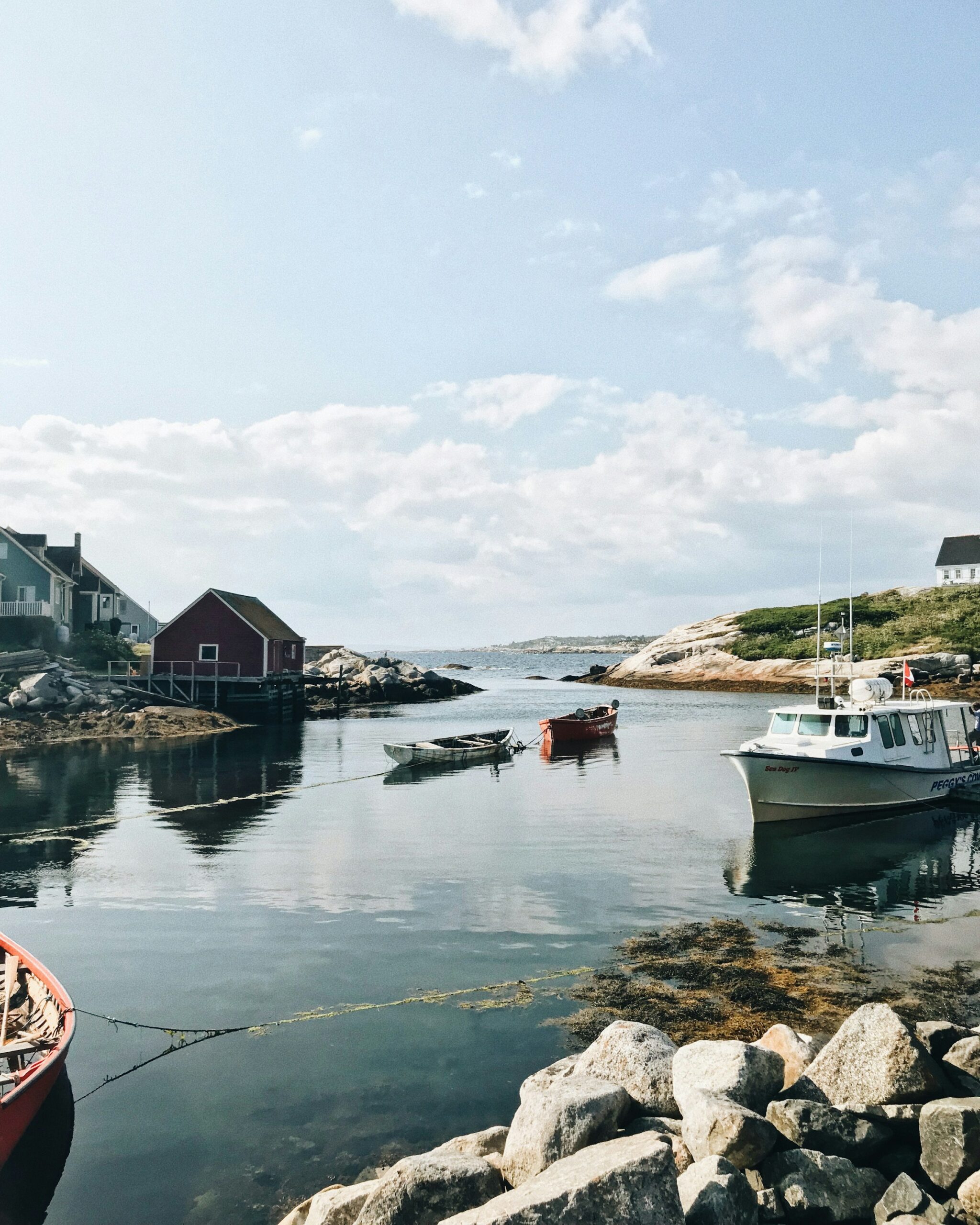 A photo from the coastline of Peggy's Cove, Nova Scotia. There are small boats in the foreground and a low mountain on the horizon line. The day is bright, with puffy clouds in the sky, and the water is calm.