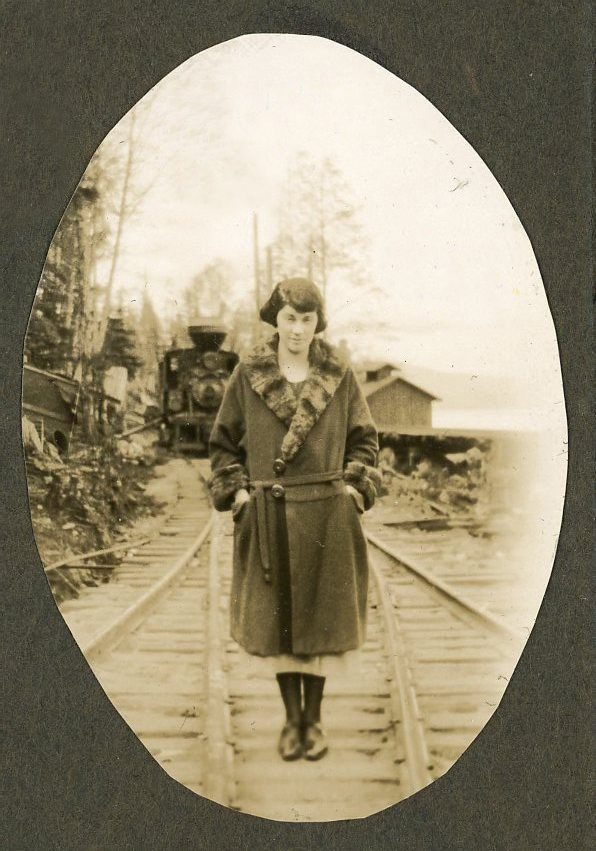 A vintage photograph of a light-skin toned woman in a fur-trimmed coat, standing on a train track. There's a stopped train at the station in the background.