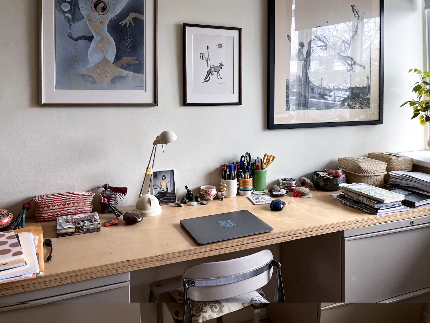 A photo of Beatriz Hausner's working space, where a long desk has a laptop and various stacked notebooks, cups for pens, and a slender work lamp. There are framed art prints on the wall and the room is softly, naturally lit by the daylight.