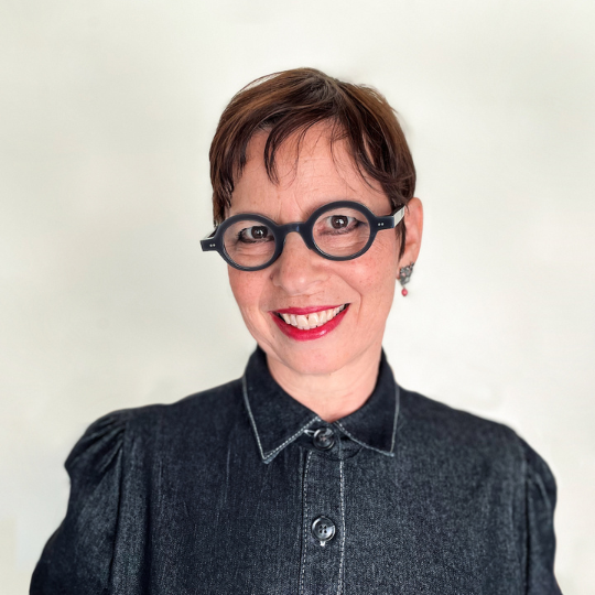 A photo of poet Beatriz Hausner. She is a light skin-toned woman with dark, short hair in a pixie cut, bold round glasses and red lipstick. She smiles widely for the camera.