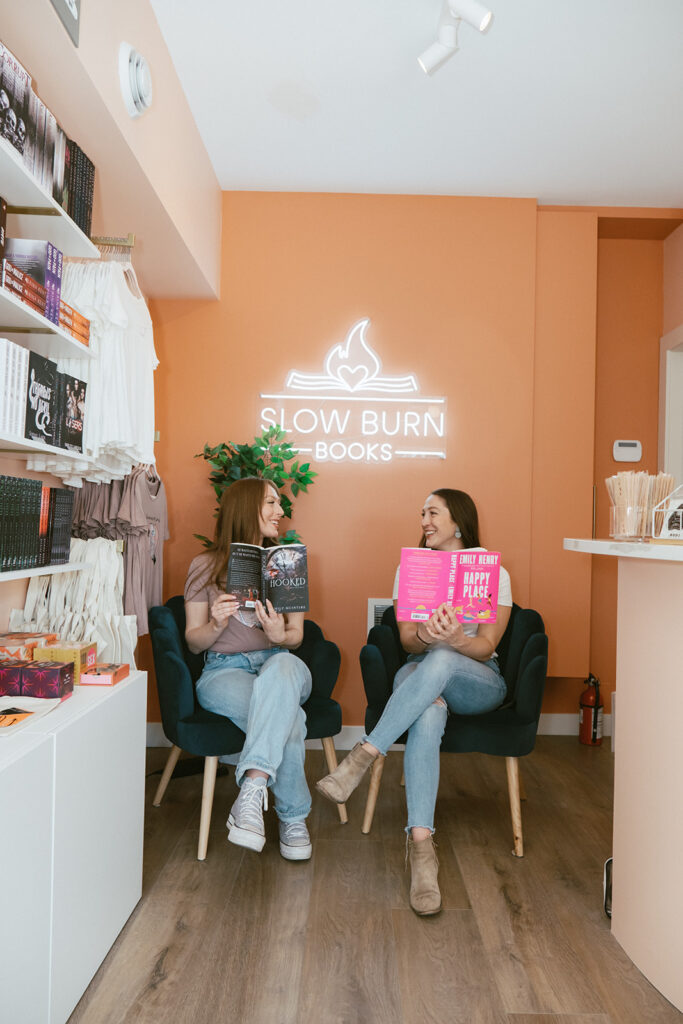 Slow Burn Books' Nicola and Shannon, holding romance novels and sitting in their shop. A neon sign with the shop's name is on the wall behind them.
