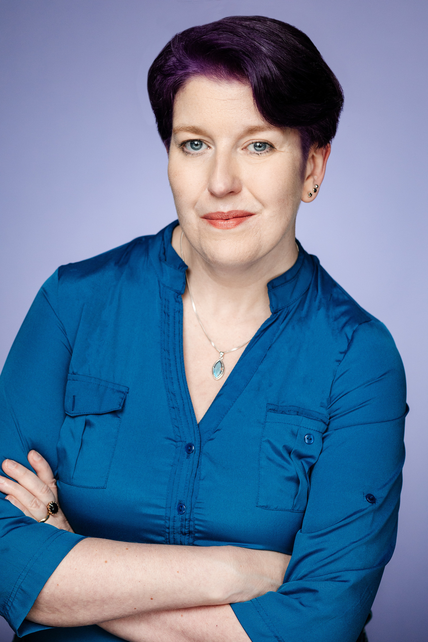 A photo of poet AJ Dolman. They are a femme-appearing, light-skin toned person with short, dark purple hair, and they stand with their arms crossed, wearing a blue button down shirt.