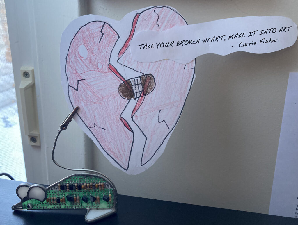 A drawing of a broken heart bandaged together is held up by a mouse-shaped photo holder. An attached quotation reads "Take your broken heart, make it into art." -Carrie Fisher