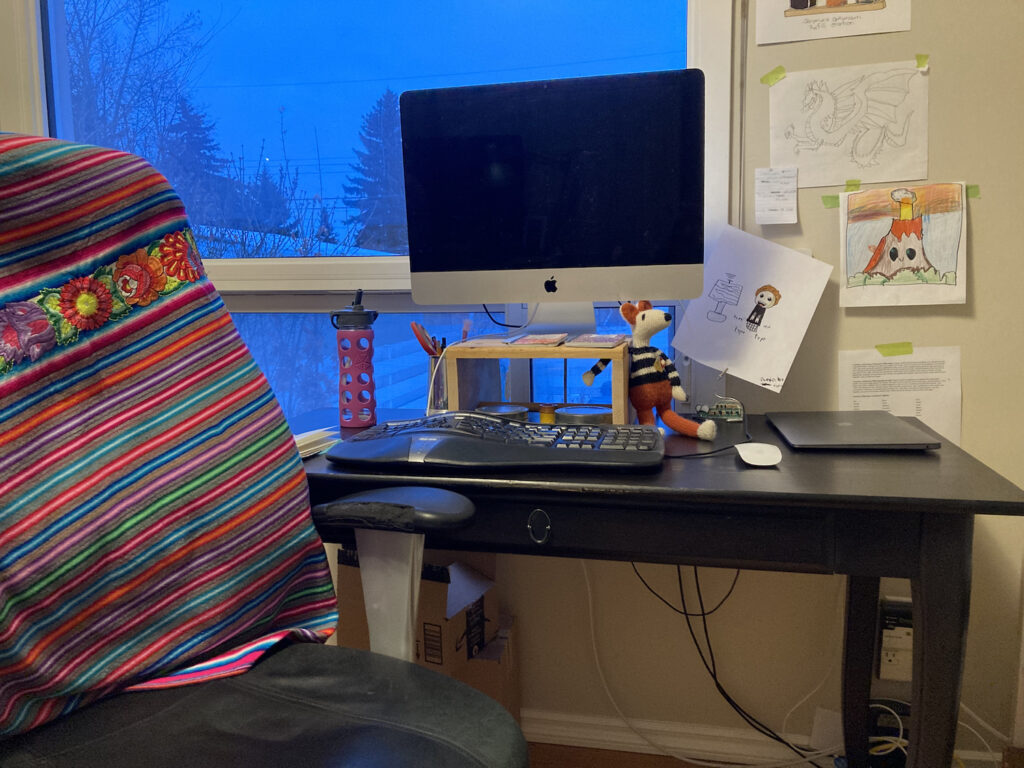A photo of Jaymie Heilman's workspace. A large Mac computer monitor stands on a riser on the dark desk, and a colourful blanket covers the back of the chair. There are drawings tacked up on the wall. The window beyond shows trees at dusk or late evening. 