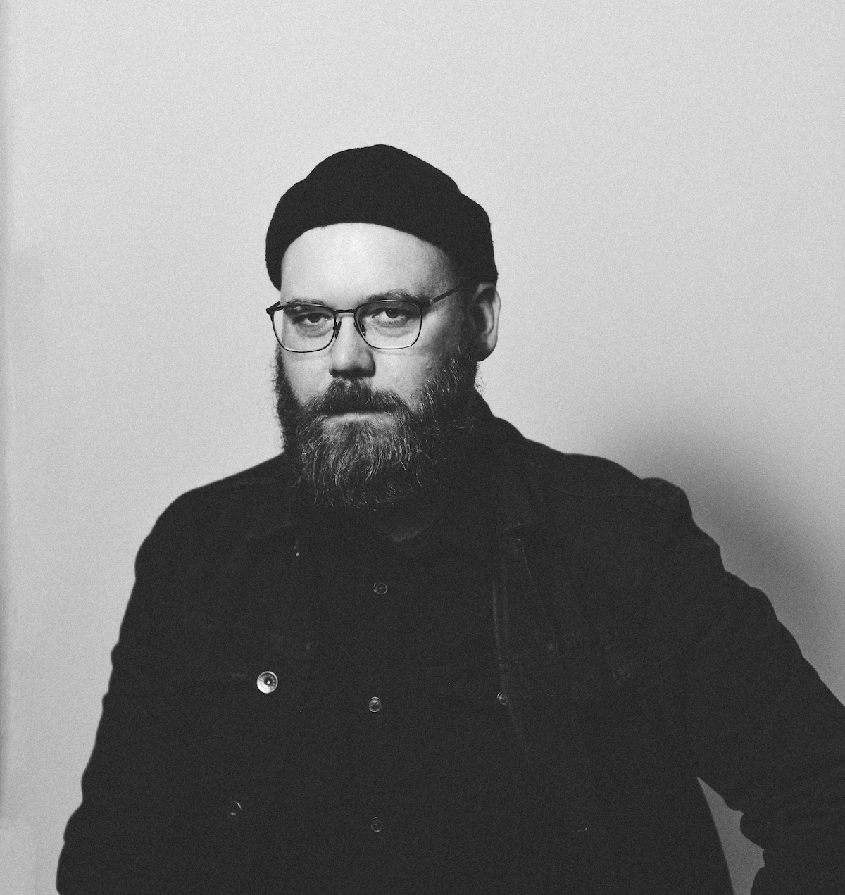A black and white photo of poet Joshua Chris Bouchard. He is a light-skin-toned man wearing a small toque and wire-rimmed glasses, and he has a thick beard.
