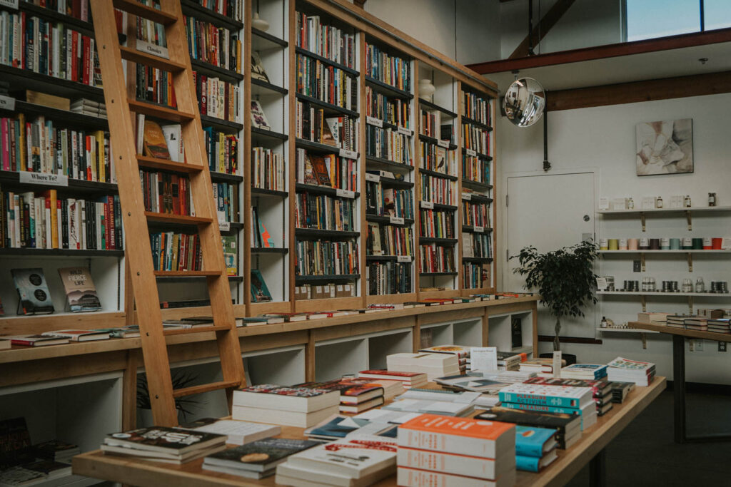 A photo of the interior of Upstart & Crow, showing tall shelves stocked with books, and a rolling ladder set across the shelves. A table in front is also stacked with books.