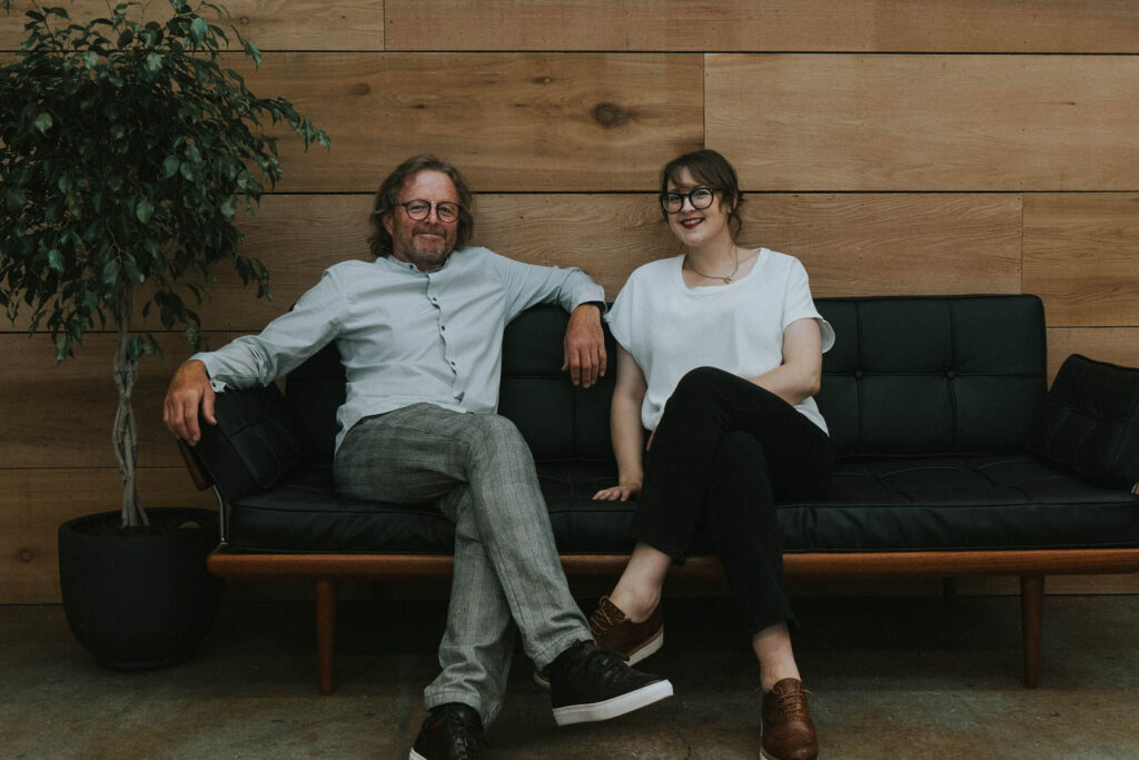 A photo of Upstart & Crow's Ian and Zoe, sitting next to each other on a sofa. 