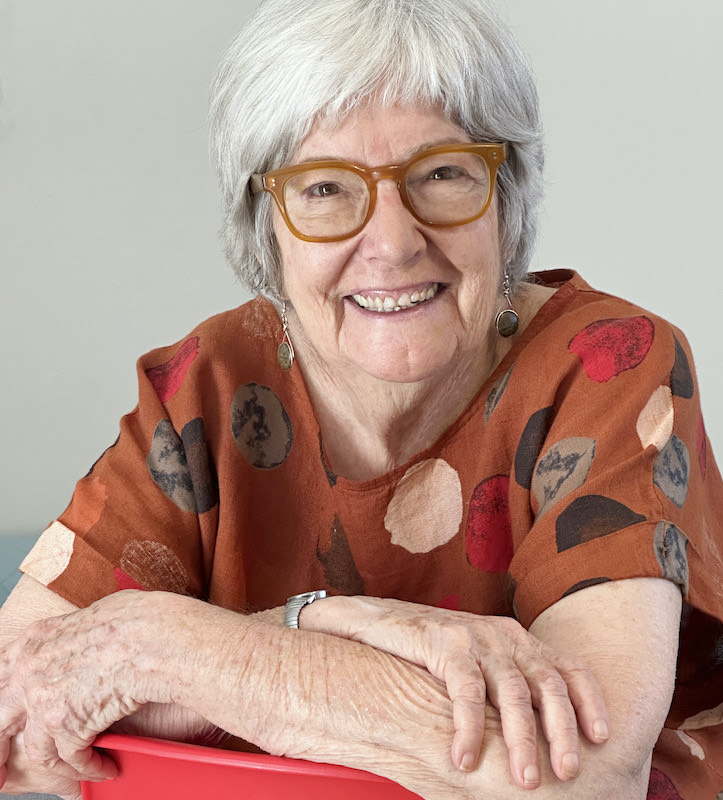 A photo of author Robin Pacific. She is a light skin-toned woman with short grey hair, wide-framed brown glasses, and an orange t-shirt. She crosses her arms and smiles at the camera.