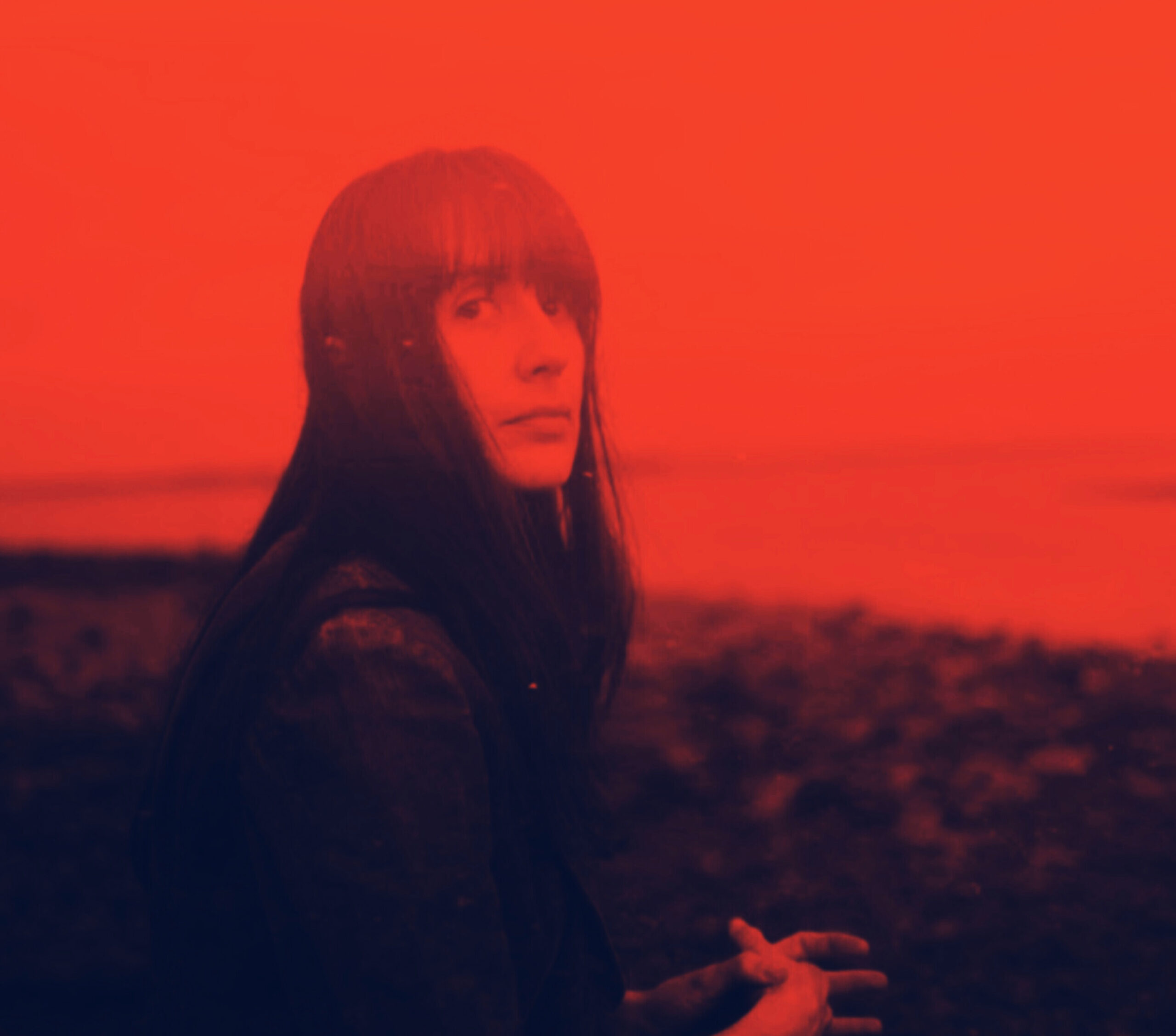 A photo of the poet Colleen Coco Collins. A red-tinted photograph, the poet stands by a shoreline, looking over her shoulder.