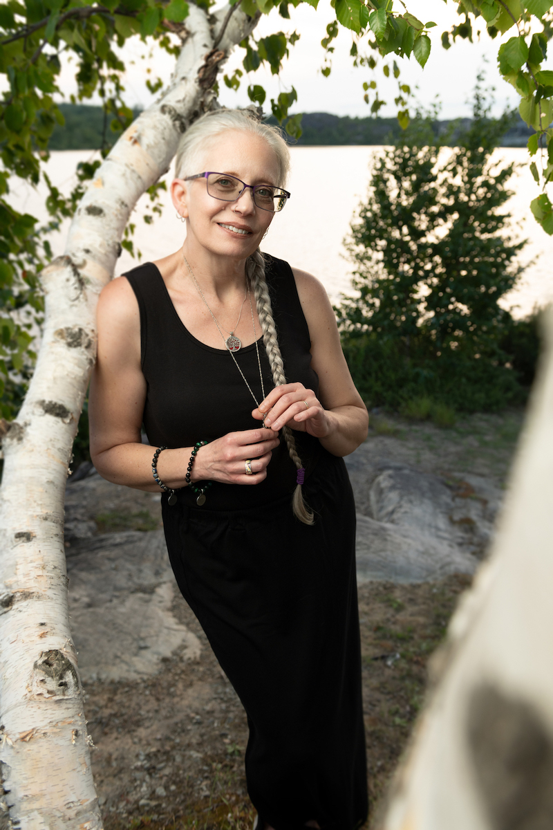 A photo of poet Melanie Marttila. A light-skin toned woman with long grey hair in a braid and glasses. She leans against a birch tree in a black sleeveless dress, with a lake in the background.