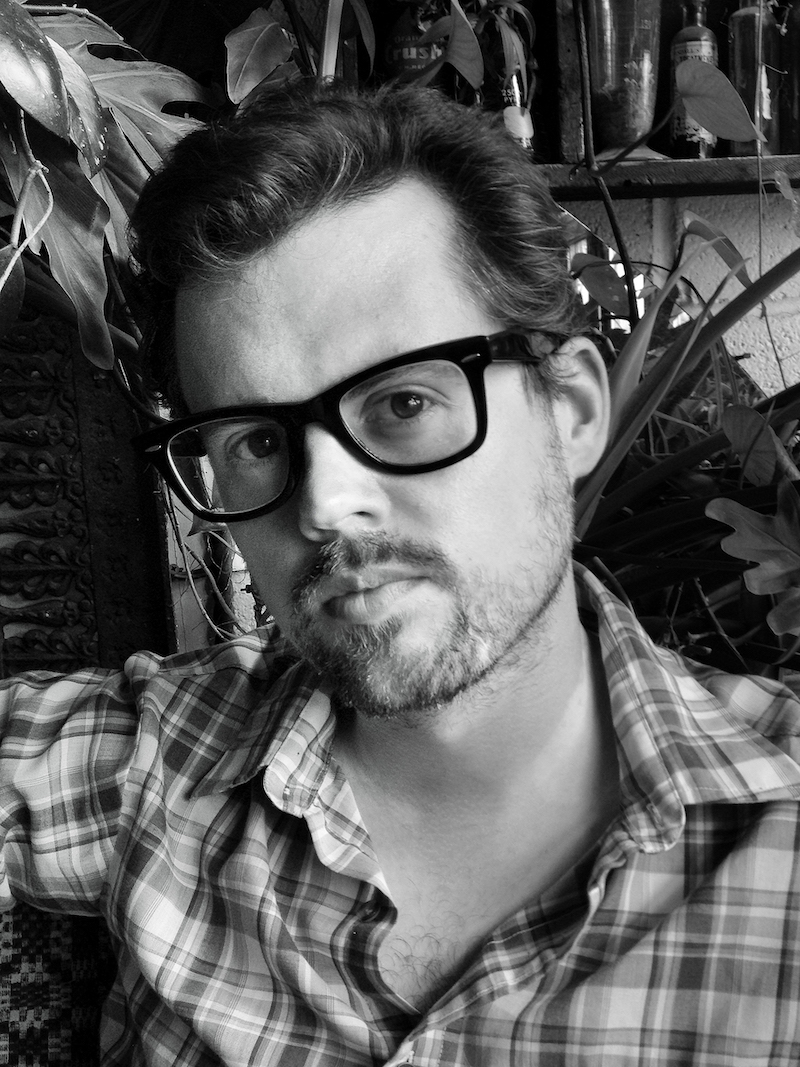 A photo of poet Jamie Sharpe. He is a light-skin toned man with dark, short hair and thick-framed glasses, wearing a plaid shirt.