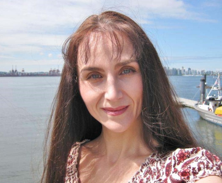 A photo of Svetlana Ischenko. She is a light skin-toned woman with straight, reddish-brown hair, standing before a body of water on a sunny day with city buildings visible on the far shore.