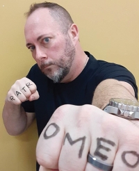 A photo of author Gerald Arthur Moore. He is a light-skin toned man with short hair and beard, and wears a black tshirt. He punches toward the camera lens, the letters "R A T R O M E O" are written on the knuckles of his hands.