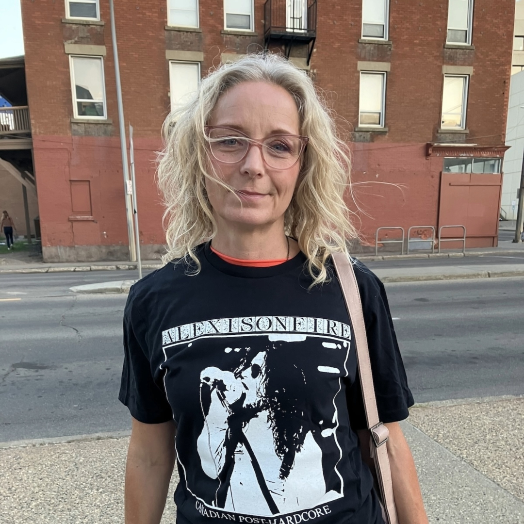 A photo of writer Robyn Braun. She is a light skin-toned woman with medium length blonde hair and glasses. She stands outside, in front of a brick building with many windows on its multi-storey facade. Robyn wears an Alexisonfire t-shirt.