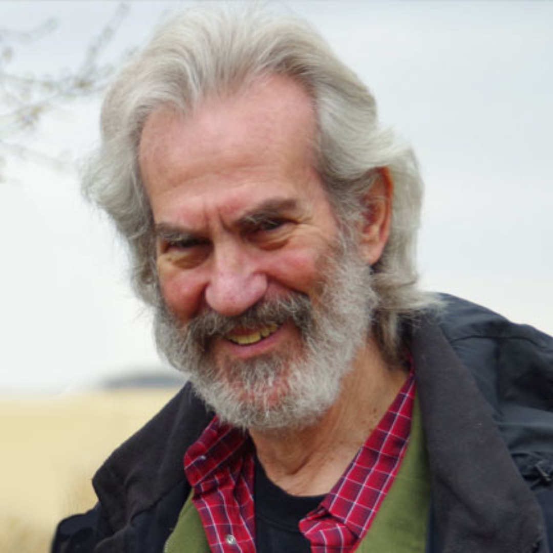 A photo of writer Dave Margoshes. He is a light skin-toned man with wavy grey hair and a grey beard. He wears a warm coat and stands in front of a prairie, blurred in the background.