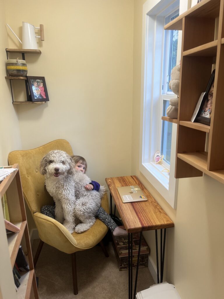 A photograph of Adrienne Gruber's writing space. A small desk with an Apple laptop on it sits before a window, with a velvet, upholstered chair at the desk. A curly-haired dog sits on a child on the chair. The child is peeking out from behind the dog's back.