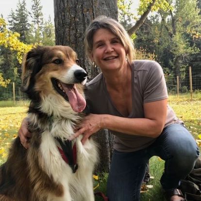 A photo of author Ellen Anderson Penno. She is a light skin-toned woman with hair pulled back into a ponytail, crouched outdoors and holding a happy-looking, large dog with its tongue lolling out of its mouth. Ellen is smiling.