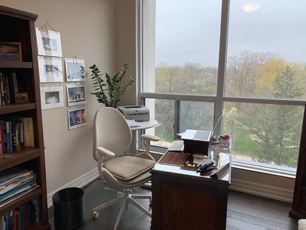 A photo of Connie Gault's office. A laptop sits perched on some risers on a wooden desk, with a modern desk chair behind it. Plants, artwork, and a full bookshelf are against the wall. Outside of the floor-to-ceiling window trees are visible.