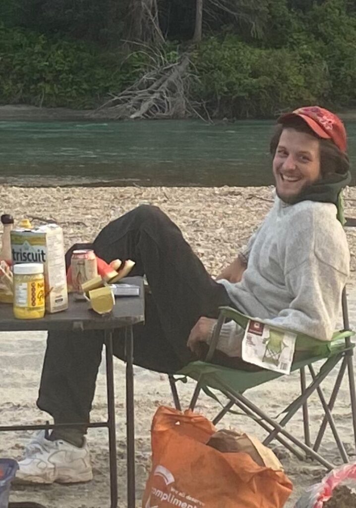 A photo of writer Hamish Ballantyne. He is a light skin-toned person in a baseball cap, sweatshirt, and track pants, sitting in a camping chair on a beach. There is a table with snack foods piled onto it. In the background, a turquoise river drifts by, with a fallen tree on its far bank.
