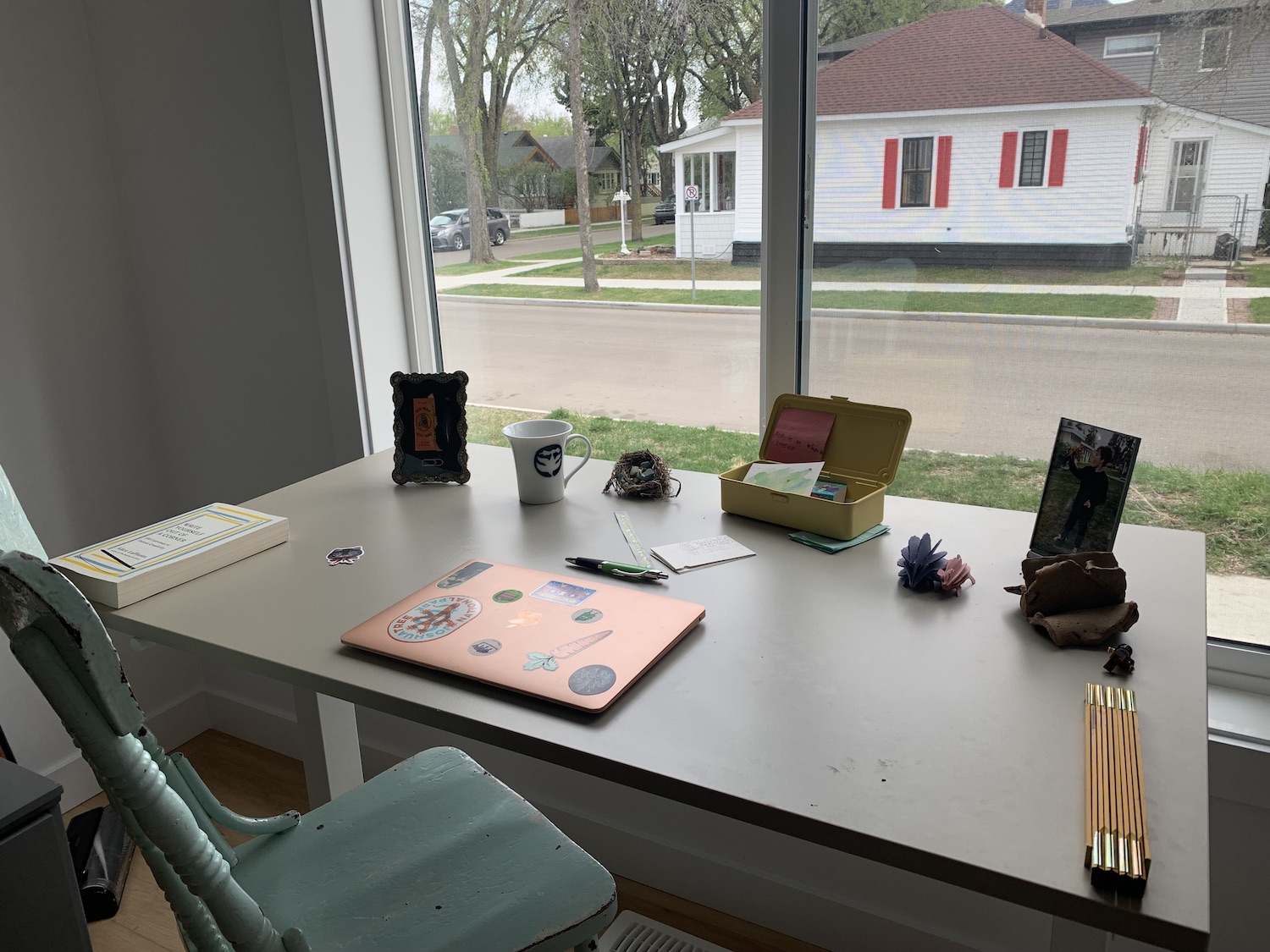 A photograph of Robyn Braun's workspace. An antique painted chair waits at a plain desk, which is scattered with photo frames, writing implements, a mug, and a closed laptop covered in stickers. A folding ruler sits in the corner. Out the window, there is a white house with red shutters visible across the street.