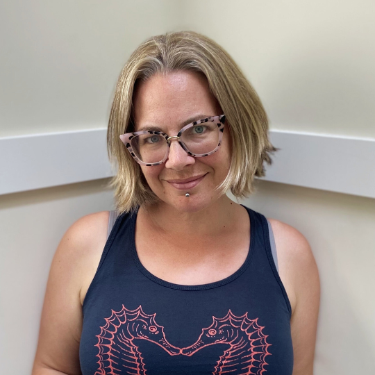 A photo of writer Adrienne Gruber. She is a light-skin toned woman with chin-length, straight blonde hair, wearing plastic-framed tortoiseshell glasses and a navy tank top with two seahorses on it.