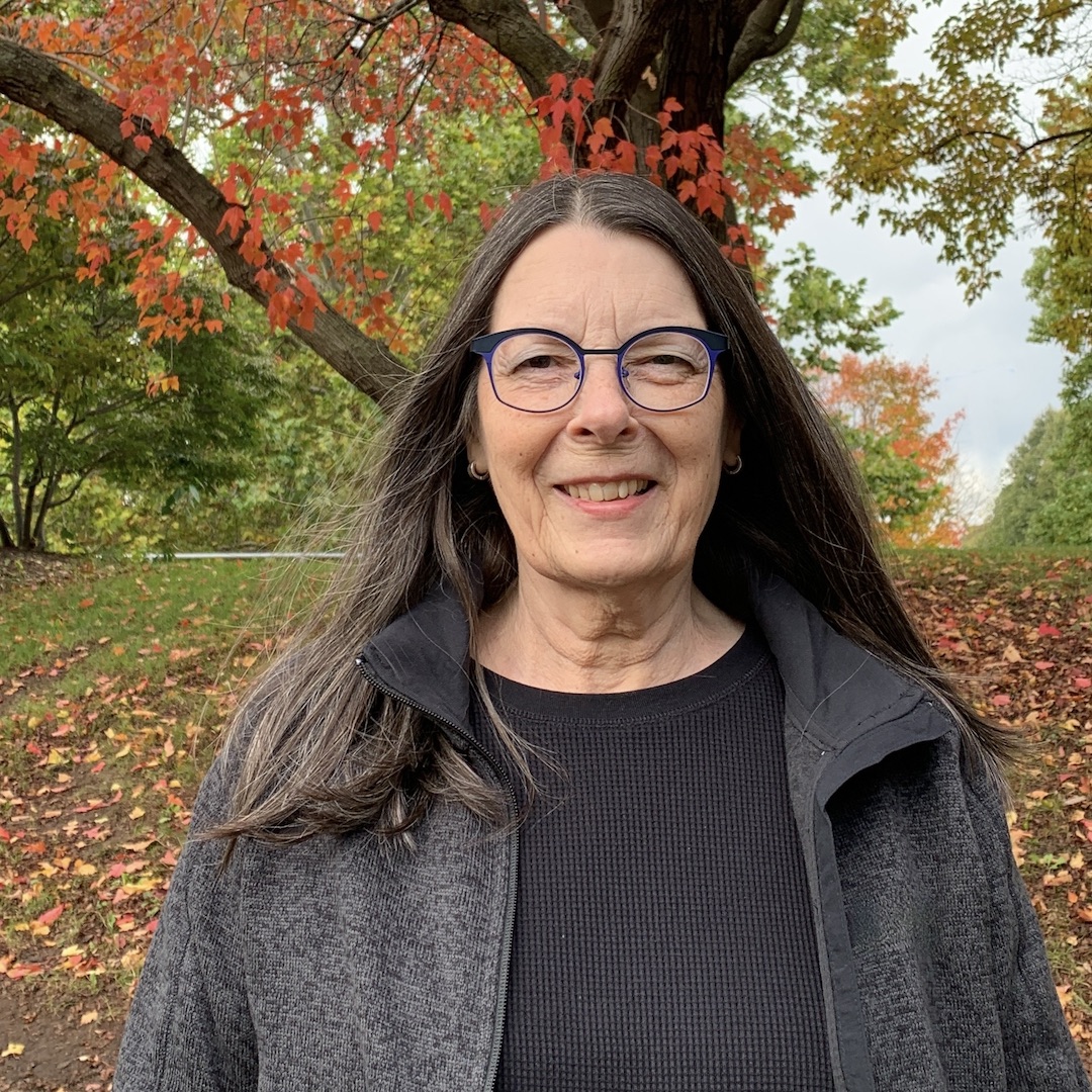A photo of writer Connie Gault. A light skin-toned woman with long dark hair, she wears glassea nd a dark jacket, and stands outside amid fall leaves.