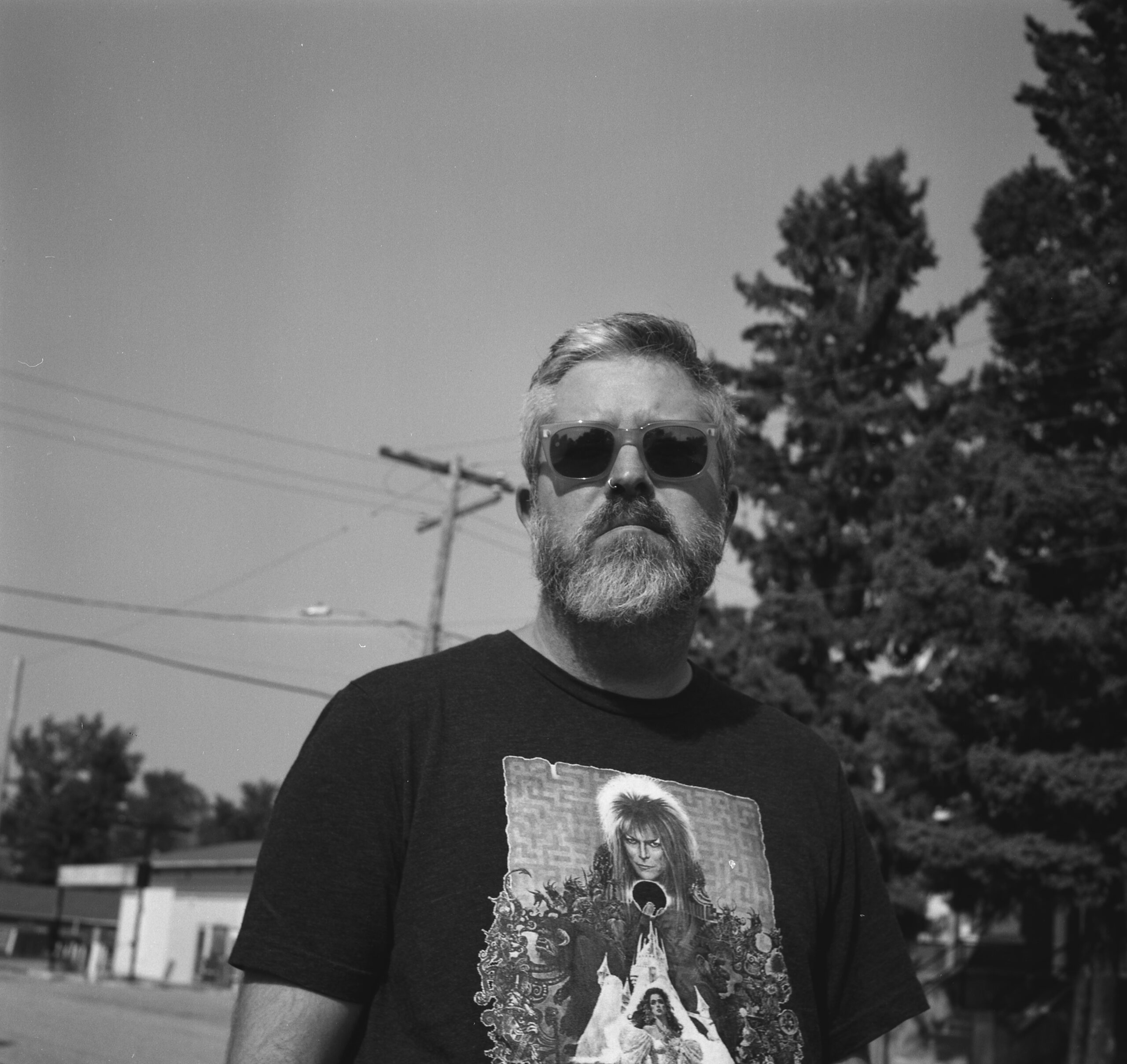 A black-and-white photo of author Jes Battis. They have facial hair and are wearing sunglasses and a graphic t-shirt of David Bowie from Labyrinth. They are standing by a road with trees and a house. 
