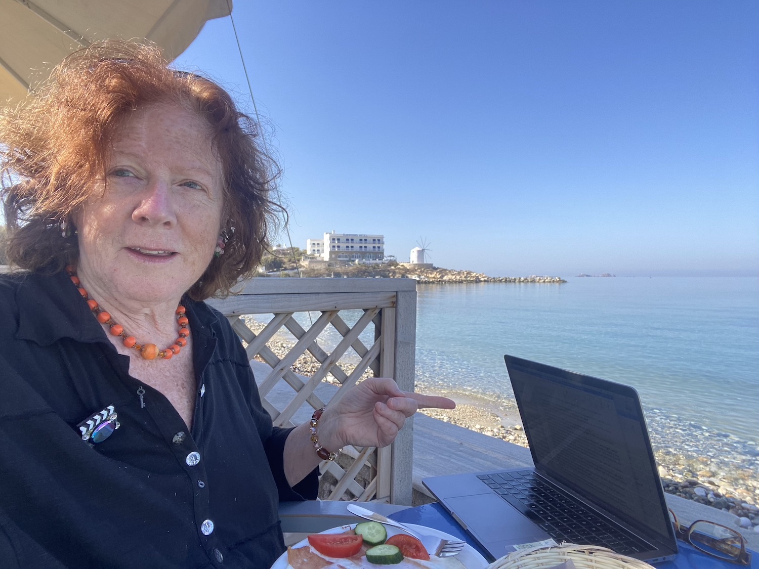A photo of Lulu Keating by the Greek coastline. She sits at a table with the remnants of a salad and her half-closed laptop, and points outward, toward the dazzling blue water.